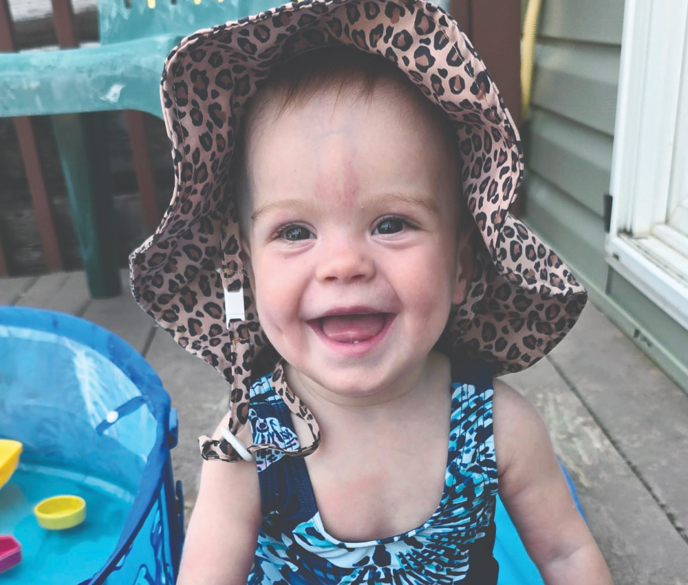 Smiling baby wearing a sun hat and swimsuit sitting in a baby pool.